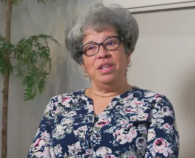 Middle-aged woman with gray curly hair and glasses wearing floral blouse sitting indoors near plants.