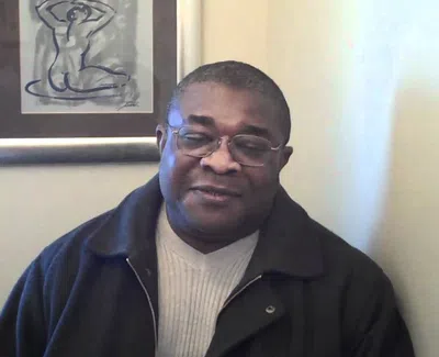 Man wearing glasses and a dark jacket sitting indoors with framed abstract art on the wall behind him