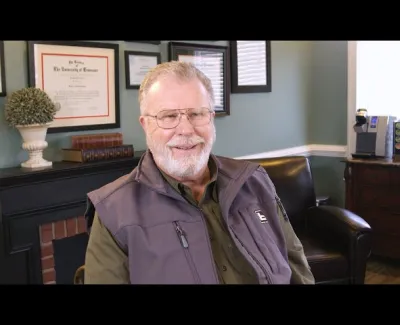 Smiling elderly man with glasses and beard sitting in office with framed diplomas and cozy decor.