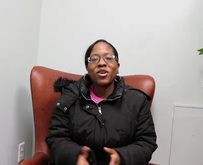 Woman in glasses and black jacket sitting in brown chair against light wall near green plant