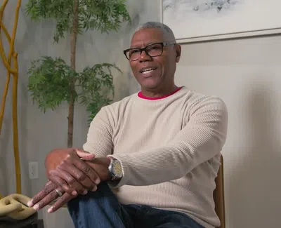 Smiling man wearing glasses and a beige sweater sitting indoors with decorative plants and wooden art pieces