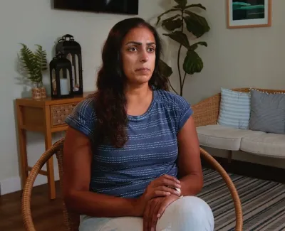 Woman with long dark hair sitting on a wicker chair in a cozy living room with plants and soft lighting.