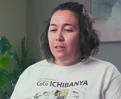 Woman wearing a CoCo Ichibanya t-shirt sitting indoors near a window with green plants.