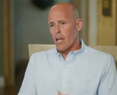 Man in light blue shirt speaking indoors with neutral background and soft lighting