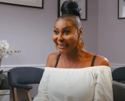 Smiling woman in a white off-shoulder blouse and large earrings sitting in a modern room with chairs and artwork.