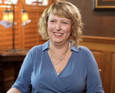 Smiling woman with short blonde hair wearing a blue blouse and necklace in a warmly lit room with wooden shutters.