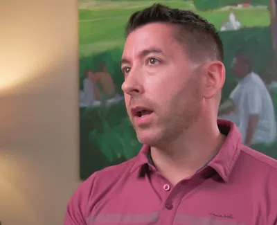 Man in a pink shirt speaking indoors with a lamp and green painting in the background