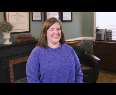 Woman smiling wearing a purple sweater sitting in a cozy office with framed certificates and dark leather chair.