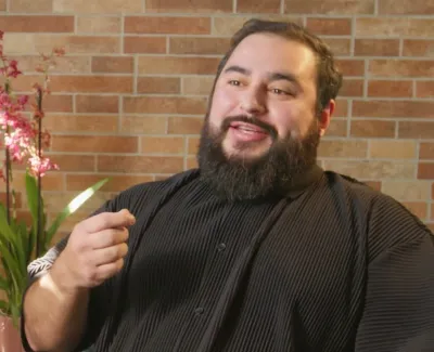Bearded man in black shirt speaking indoors with brick wall and green plants in the background