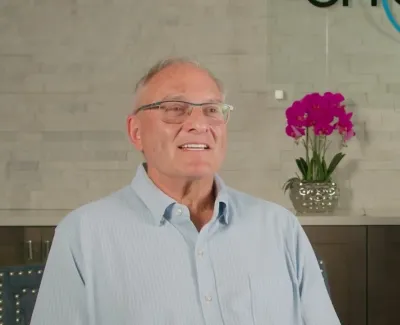 Smiling elderly man wearing glasses and light blue shirt sitting indoors with a pink orchid and stone wall background.