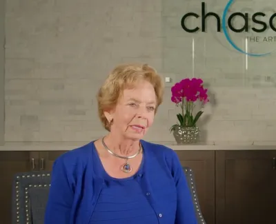 Mature woman in blue shirt sitting in dental office with Chasolen Dentistry logo and purple flowers behind her.
