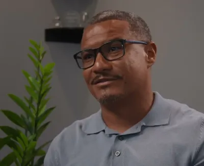 Man wearing glasses and a gray polo shirt smiling indoors with green plants and awards in the background