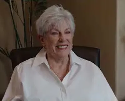 Smiling elderly woman with short white hair wearing a white shirt sitting in a cozy room with plants.