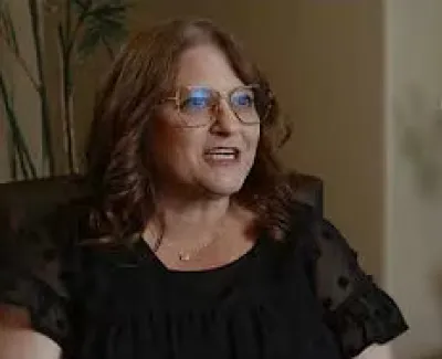 Middle-aged woman with glasses and curly hair speaking, sitting indoors near a white orchid plant.