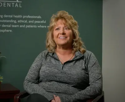 Smiling woman in gray sweater sitting in dental office with flowers and plaque on table behind her.