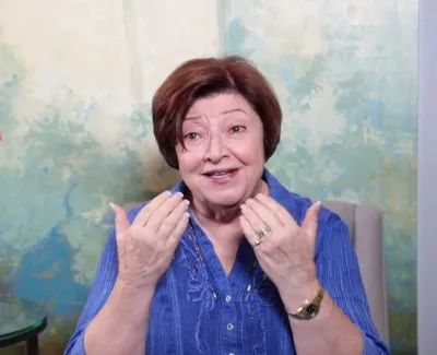 Middle-aged woman in blue shirt gesturing with hands while sitting next to a table with a vase of orange roses