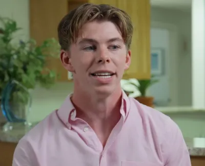 Young man in a pink shirt speaking indoors with a kitchen background and plants visible.