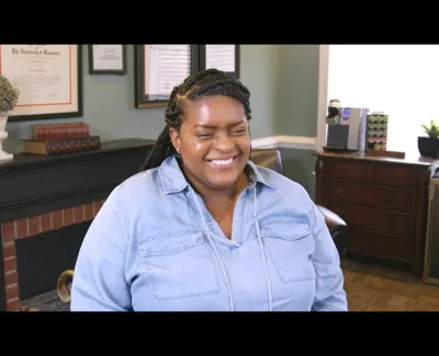 Smiling woman with braided hair wearing a blue shirt sitting in a cozy office with framed certificates and wooden furniture.