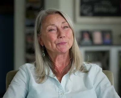 Smiling mature woman with long gray hair wearing a light blue shirt sitting in a cozy home setting.