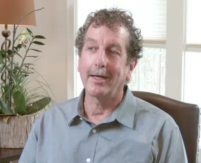 Middle-aged man with curly hair and mustache sitting in a chair in a living room with plants and a lamp.