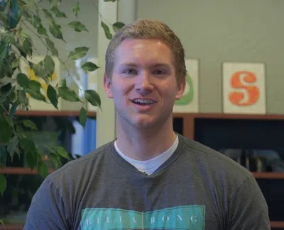 Young man in a Billabong shirt smiling indoors with bookshelf and plant in the background