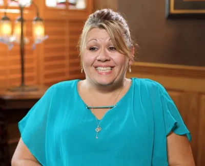 Smiling woman with blonde hair wearing turquoise blouse in warmly lit wooden room with magazines and lamp.