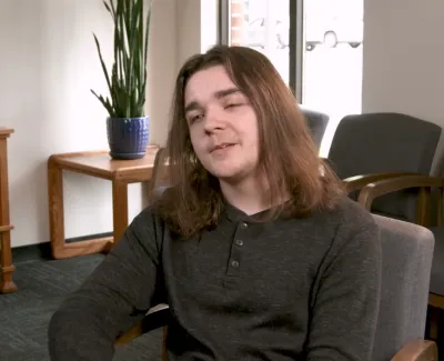 Young man with shoulder-length hair sitting in an office chair in a room with wooden furniture and a potted plant.