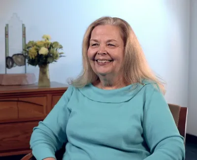 Smiling older woman in blue sweater seated in a room with wooden dresser and flower arrangements.