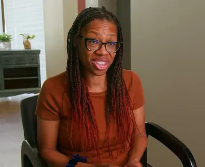 Smiling woman with braided hair and glasses sitting in a chair in a cozy home interior.