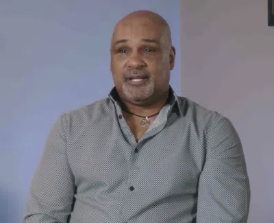 Middle-aged man with a bald head and goatee sitting in an indoor setting with light blue walls and a plant.
