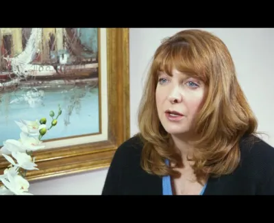 Woman with red hair speaking beside a white orchid and a framed painting of boats on water.