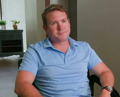 Man in light blue polo shirt sitting on chair indoors with arms resting, looking thoughtful.