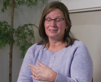 Woman with glasses in a purple sweater speaking with hand gesture in a room with indoor plants.
