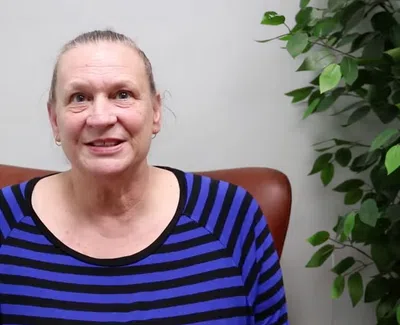 Smiling woman in a blue and black striped shirt sitting on a brown chair next to a green leafy plant