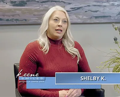 Woman with blonde hair in red sweater speaking in an indoor setting with plant and painting background.