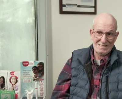 Smiling elderly man wearing glasses and a vest sitting in a dental office with toothpaste displays and services board.