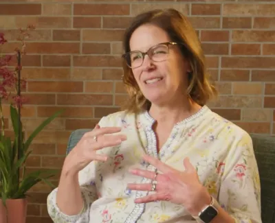Middle-aged woman with glasses speaking indoors, gesturing with hands, plants and brick wall background