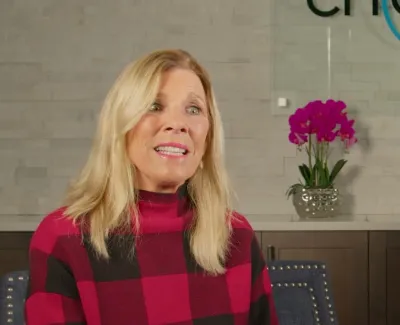 Smiling woman in red and black checkered sweater sitting in a modern dental office with orchid and logo in background.