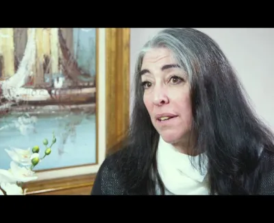 Woman with long dark and gray hair speaking in front of a framed painting of sailboats and a white orchid flower.
