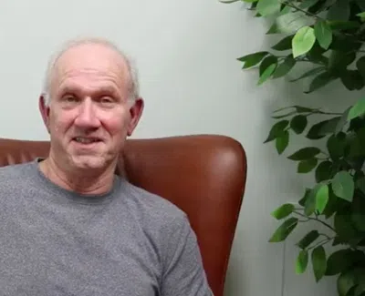 Smiling elderly man in a gray shirt sitting on a brown chair with green leafy plant nearby indoors.