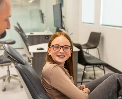 Smiling girl with braces sitting in dental chair talking to dentist in bright orthodontic office.