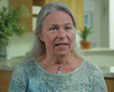 Middle-aged woman with long gray hair speaking indoors in a well-lit room with plants and kitchen background.