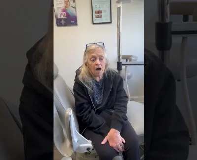 Elderly woman sitting in a medical office chair looking tired or unwell, with medical posters on the wall.