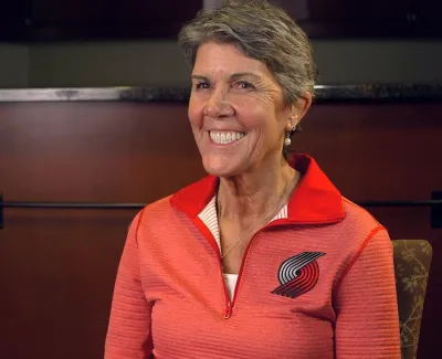 Smiling woman in a red zip-up jacket featuring a basketball team logo sitting indoors against a dark background