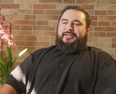 Bearded man in black shirt sitting indoors with houseplants and brick wall background, smiling and talking.