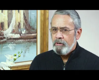 Mature man with gray beard and glasses wearing a black sweater, with a sailing ship painting and white flowers behind him.
