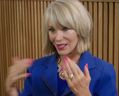 Blonde woman in blue blazer gestures with pink nails against wooden panel background and white orchids.