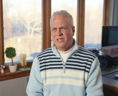 Older man in a striped sweater sitting in a bright office with large windows and indoor plants.