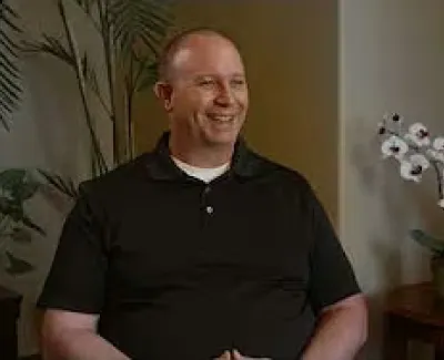 Smiling man in a black polo shirt sitting in a cozy room with plants and a table with flowers and a magazine.