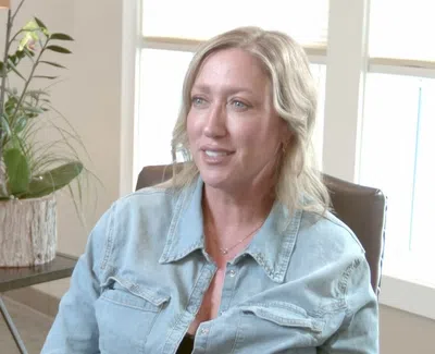 Blonde woman in a denim shirt sitting in a chair with a table of potted plants in a bright room.